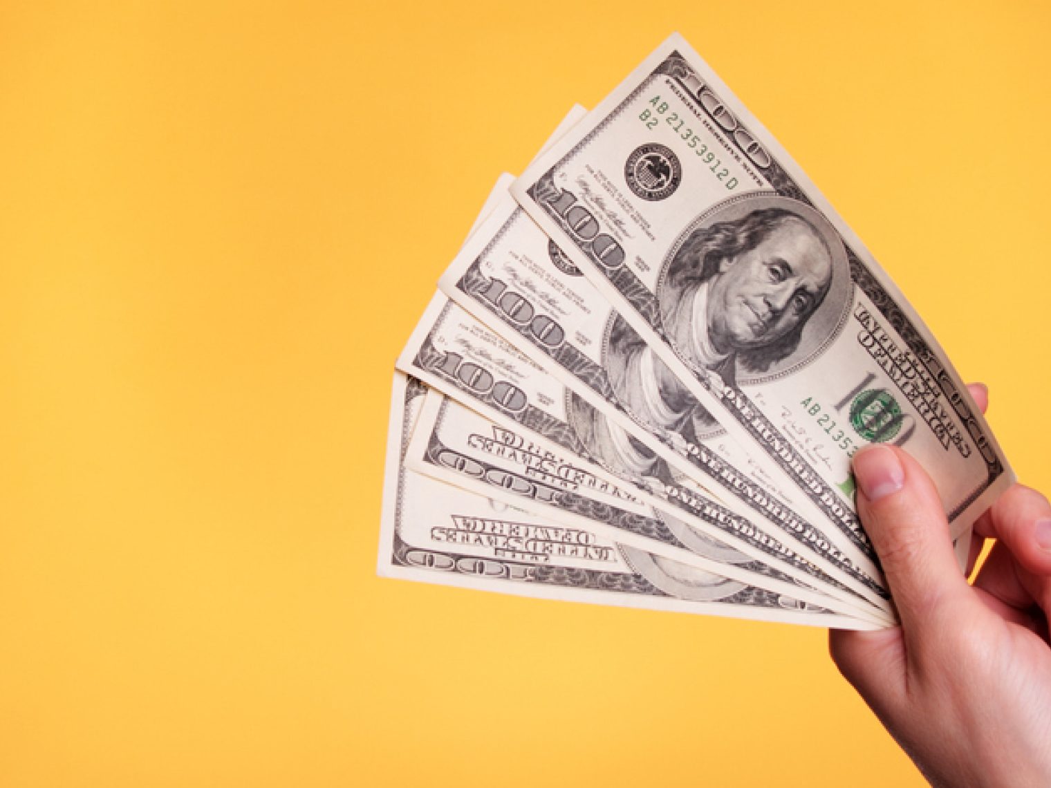 A hand holding a fanned out stack of hundred dollar bills against a bright yellow background.