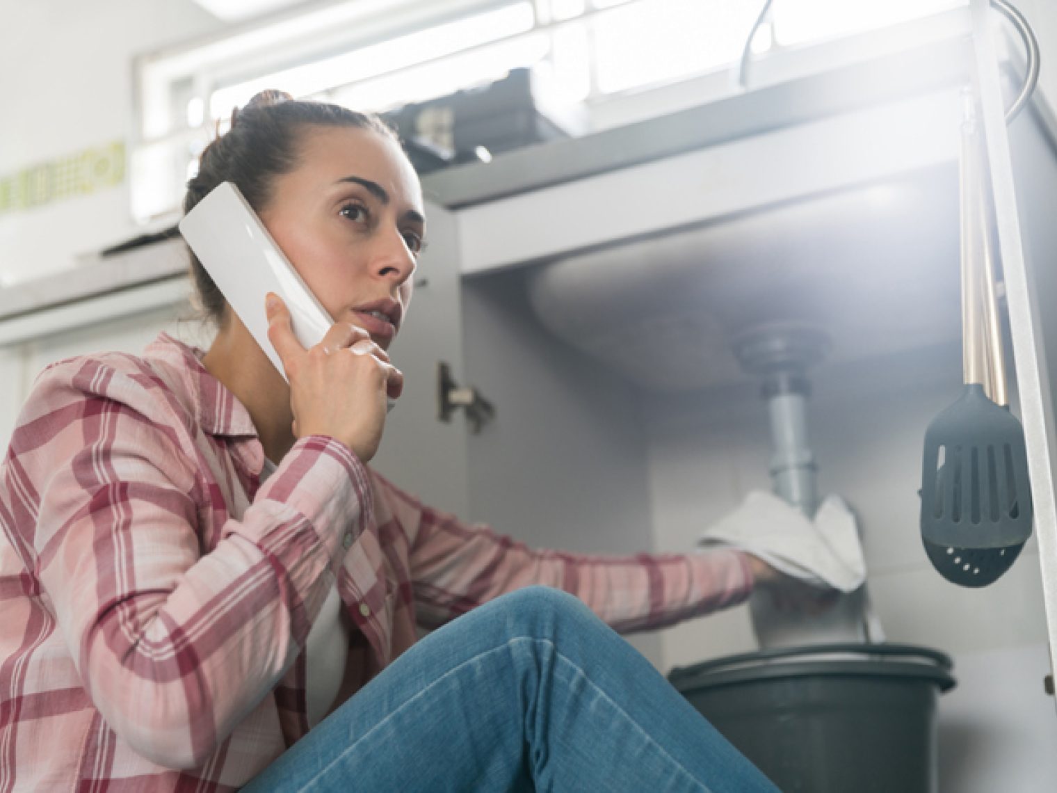 A concerned woman in a plaid shirt is talking on the phone while looking at a leaking sink with a bucket placed underneath.