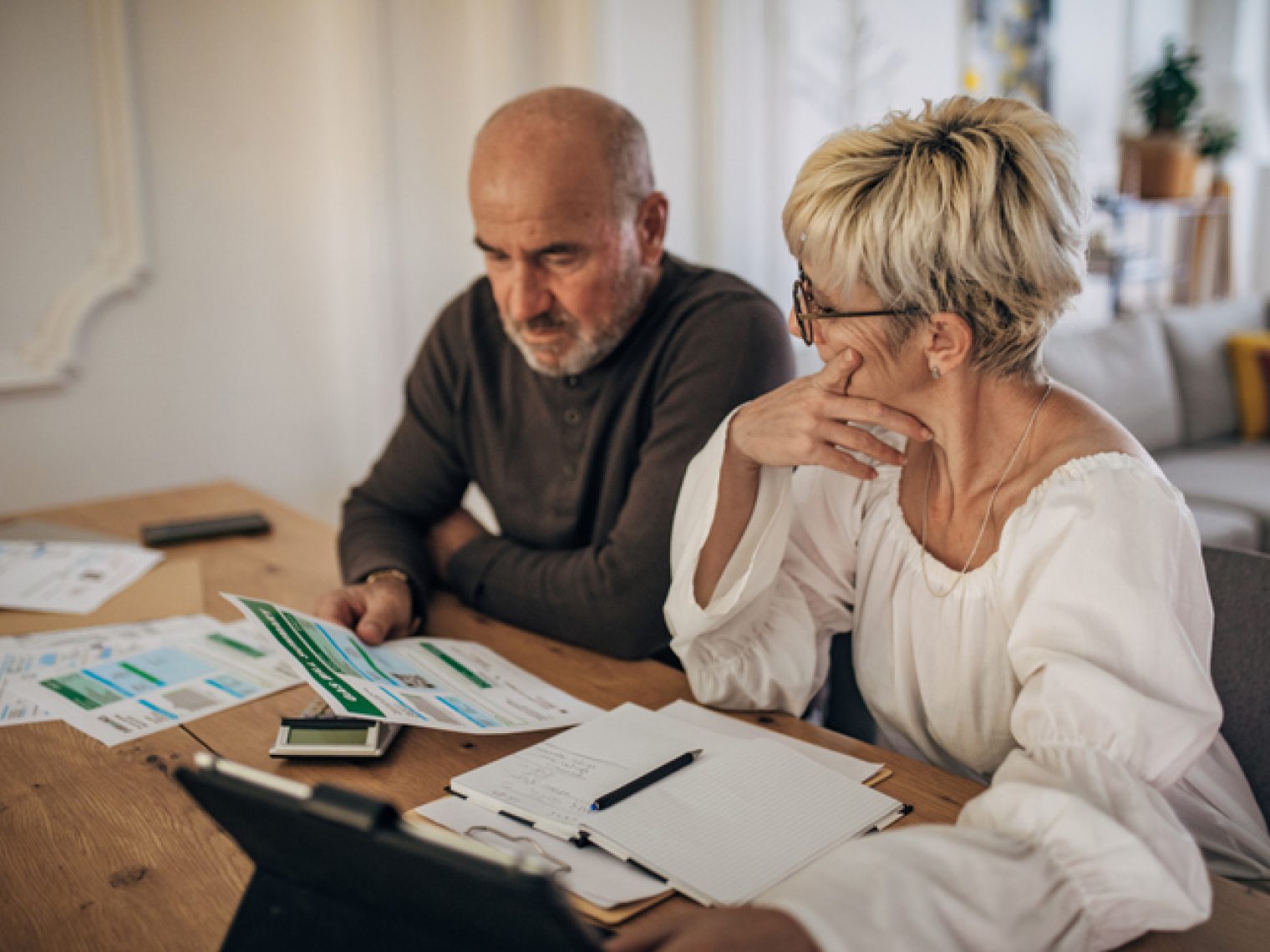 An older couple appears concerned while reviewing financial documents spread out on a table.