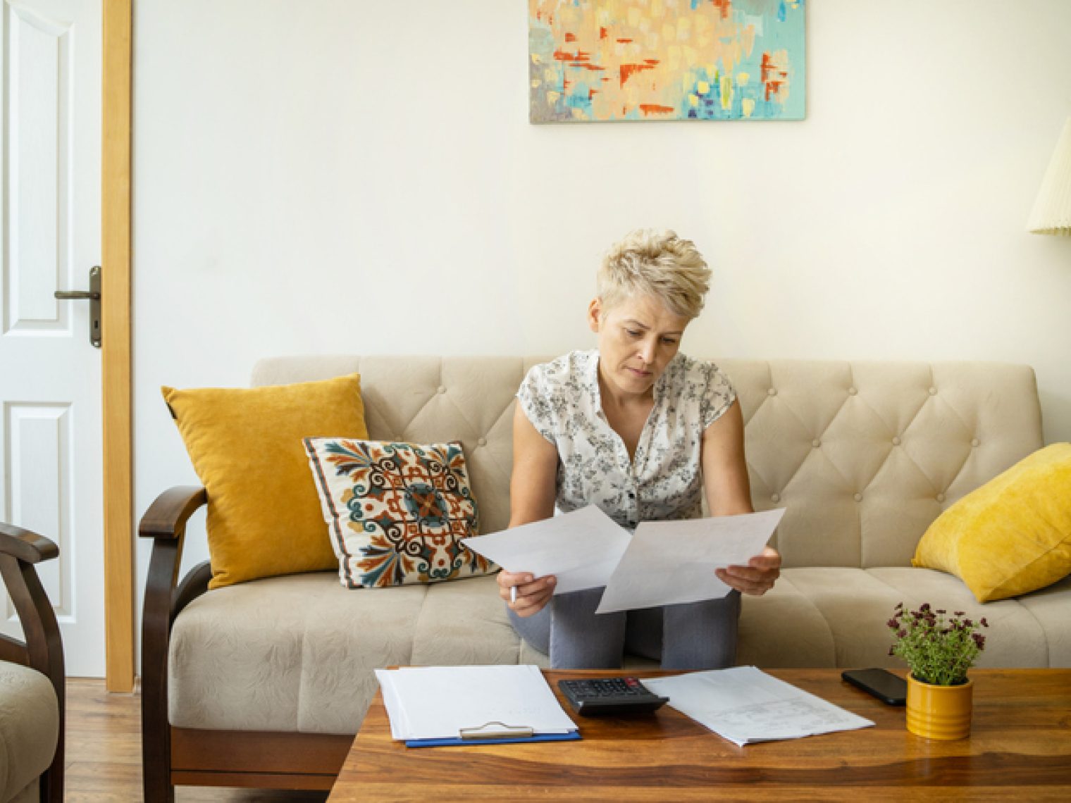 A woman with short blonde hair is sitting on a beige couch, intently reviewing two papers in her hands. She has a focused expression, suggesting she is concentrating on the content of the documents.