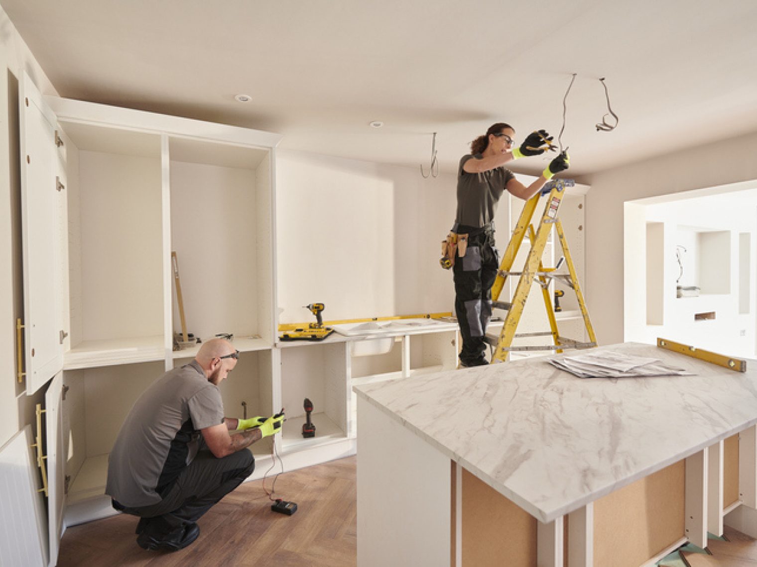 Two people are working on installing cabinets and lighting fixtures in a bright, modern kitchen, with tools and parts arranged around the room.