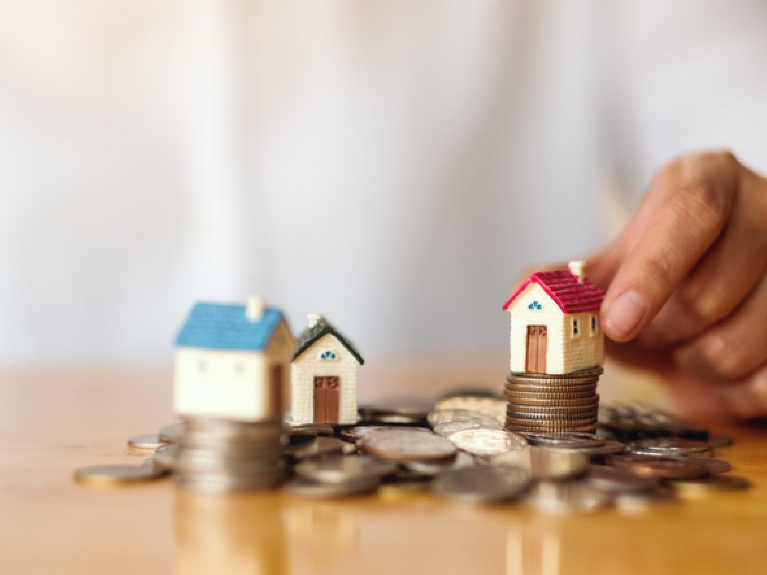 A hand is shown placing small model houses on top of stacked coins.