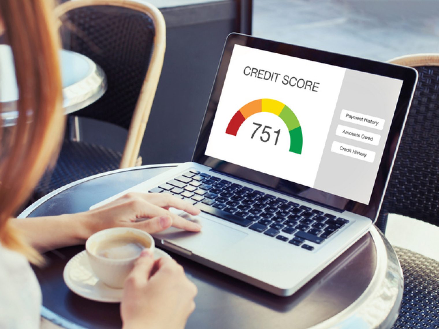 A person is sitting at an outdoor cafe table viewing a laptop screen that displays a credit score of 751, along with credit score components like payment history, amounts owed, and credit history.