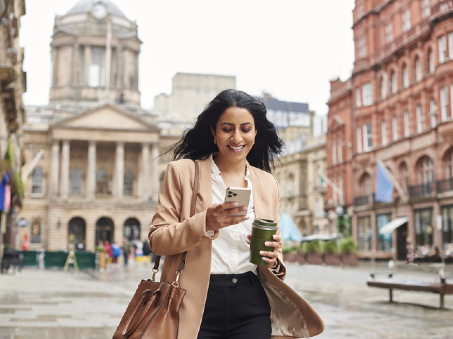 A cheerful woman, walking with a coffee cup and smartphone in hand, looks at her phone screen while walking through an urban square with historic buildings in the background.