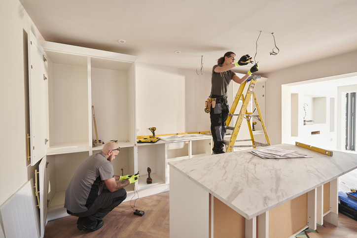 Two people are working on installing cabinets and lighting fixtures in a bright, modern kitchen, with tools and parts arranged around the room.