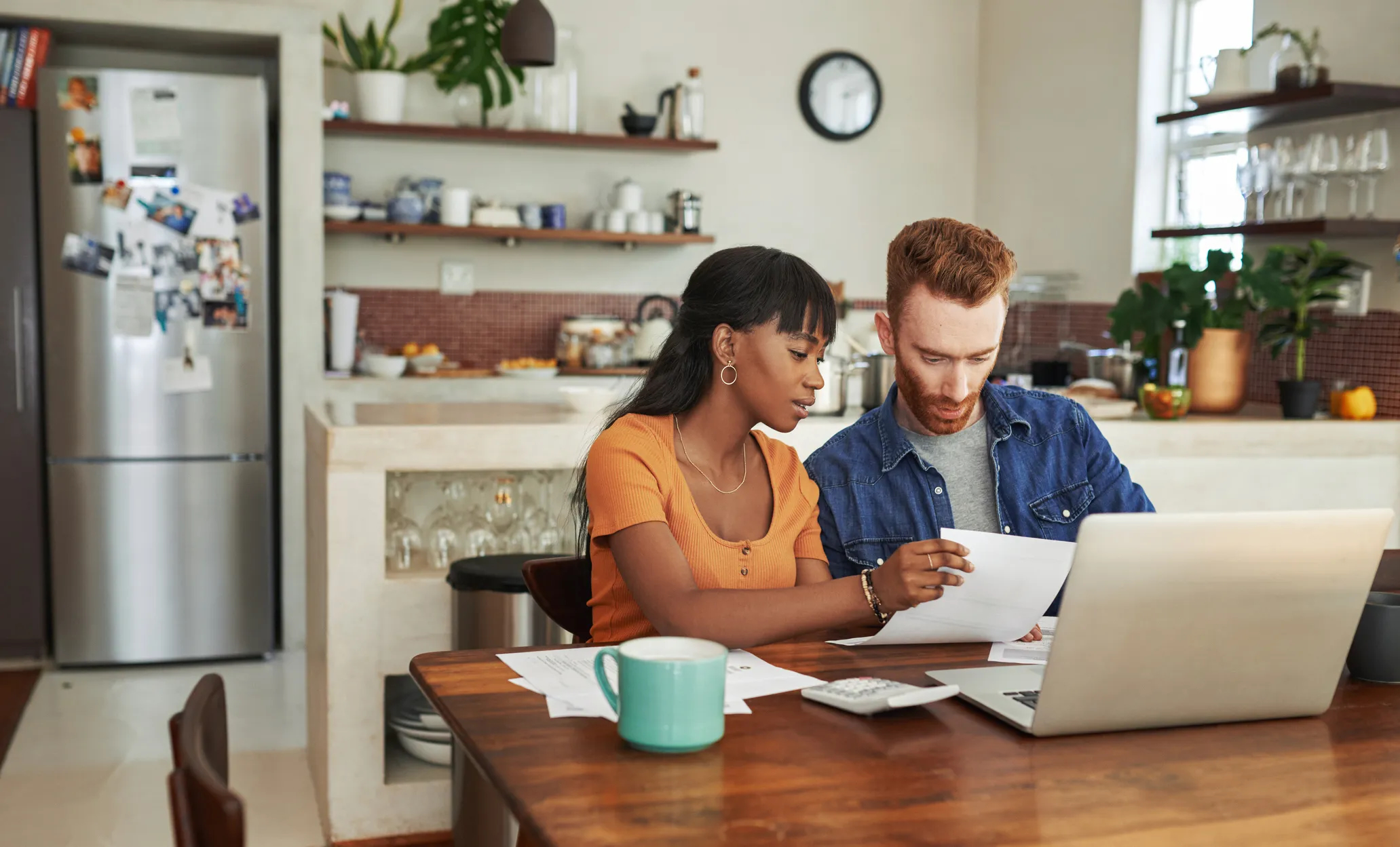 A man and woman sit at a kitchen table looking intently at a document together, with a laptop open in front of them.