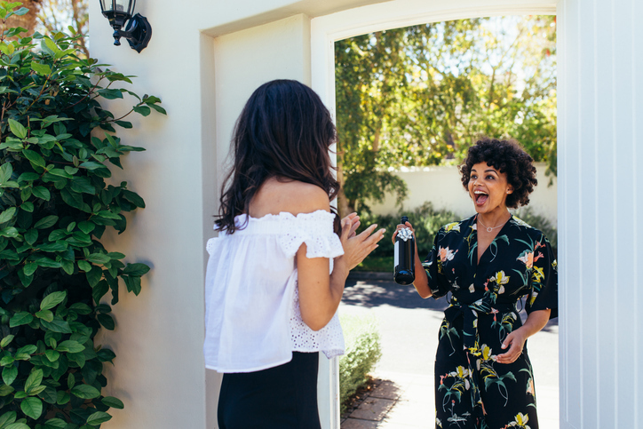 A woman in a floral dress excitedly presents a bottle to another woman at the doorway as a housewarming gift.