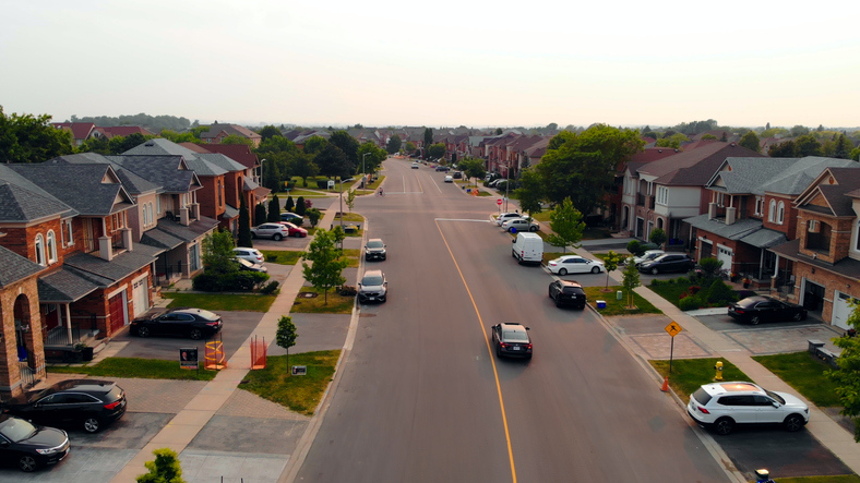 An aerial view of a residential neighborhood street with rows of detached houses, parked cars, and some traffic, captured on a hazy day.