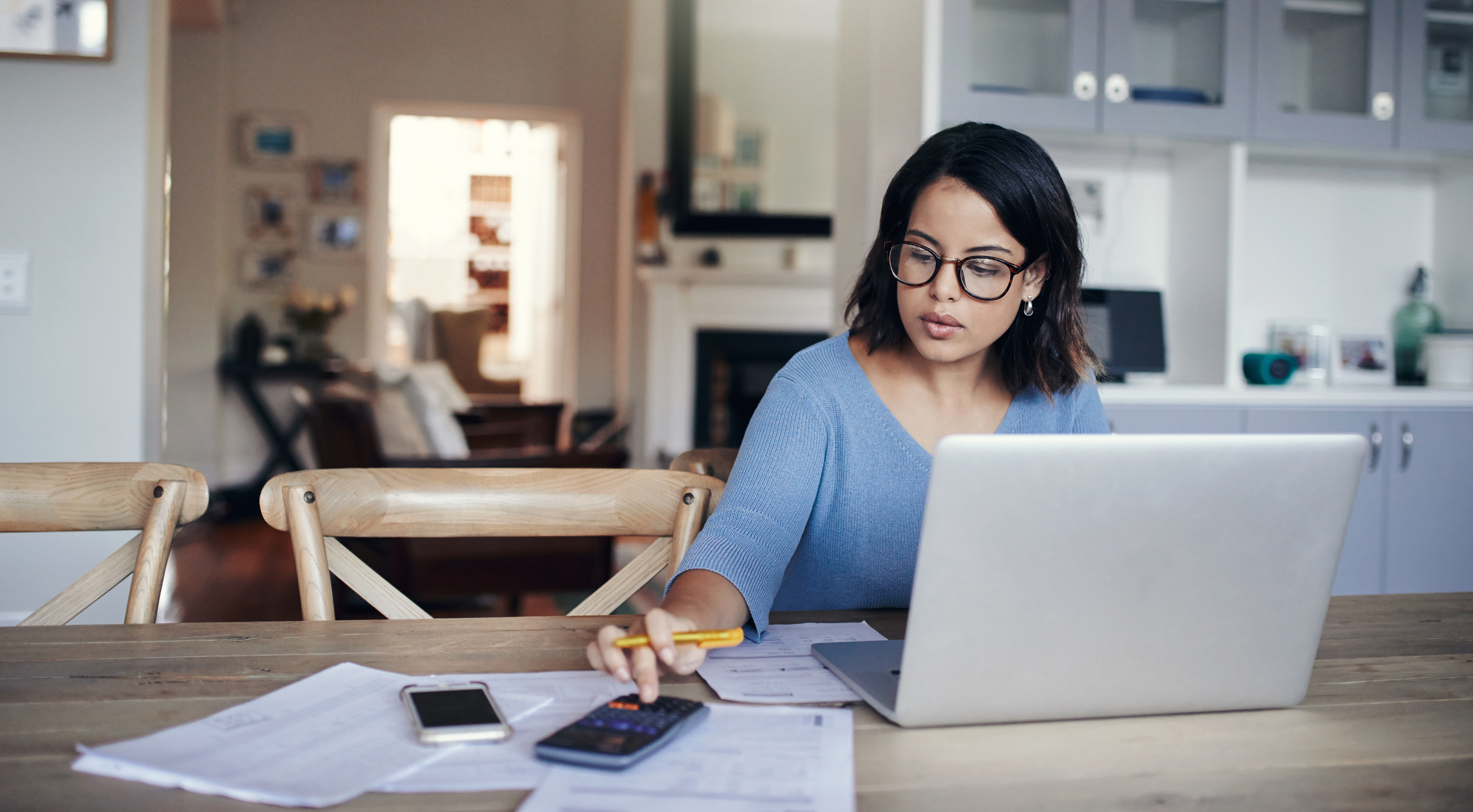 Focused woman wearing glasses and a blue sweater working at a wooden table with a laptop, calculator, papers, and a smartphone, in a cozy home setting.